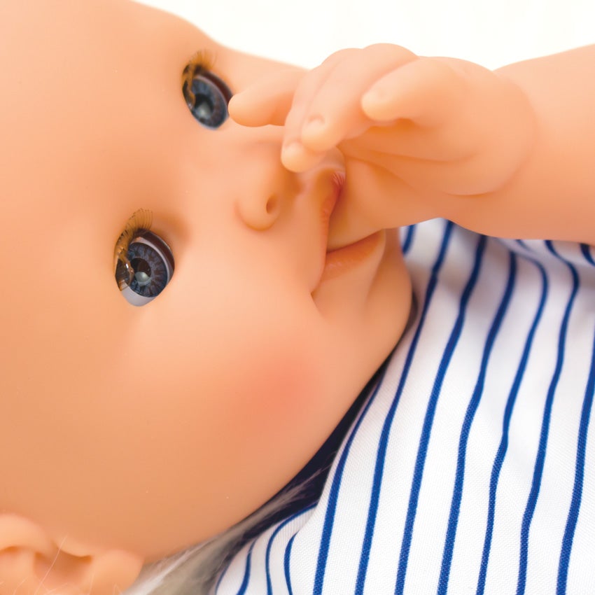 A close up shot of Corolle’s Bebe Marin’s head. The doll’s arm has been placed, so he is sucking his thumb. He is light skinned with blue, blinkable eyes. He is wearing a white with blue stripes.