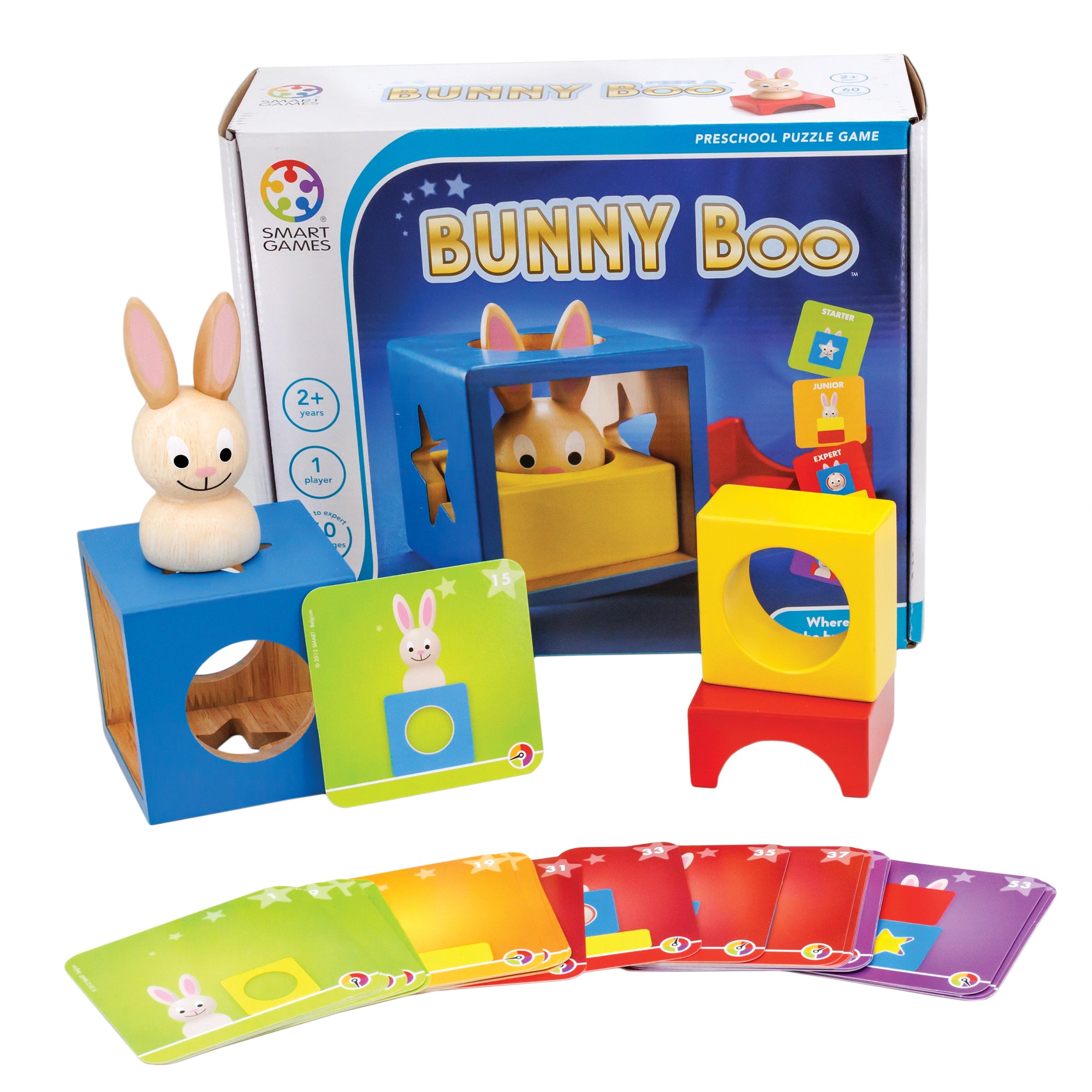 Bunny Boo game box with contents. In front of the box, is a hollow blue wood block with shapes cut out on the sides with a wooden bunny piece on top and a challenge card propped up against it. To the right are a yellow piece with a hollow circle through the middle and a red piece that shows a half circle piece cut out of the middle. There are challenge cards fanned out along the bottom in many colors.