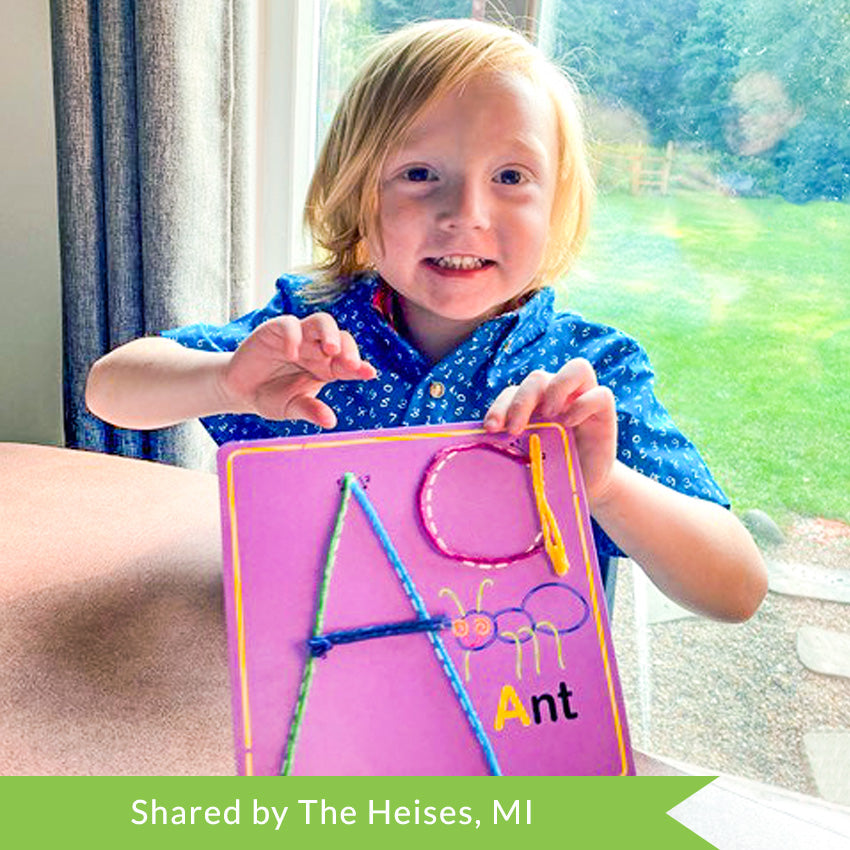 A customer photo of a young strawberry-blonde boy smiling and holding up the letter A Wikki Stick Alphabet board. The board is purple with an ant on it and the upper and lowercase letter A. He used green, light blue, dark blue, purple, and yellow wax sticks to trace the letter.