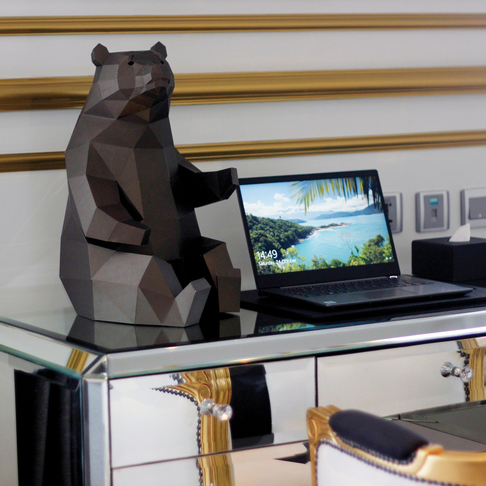 Dark brown Geometrical-shaped Papercraft bear that is folded and cut paper pieces glued together sitting on top of a mirrored desk. Next to the bear is a laptop and tissue box in front of a white wall with gold trim.