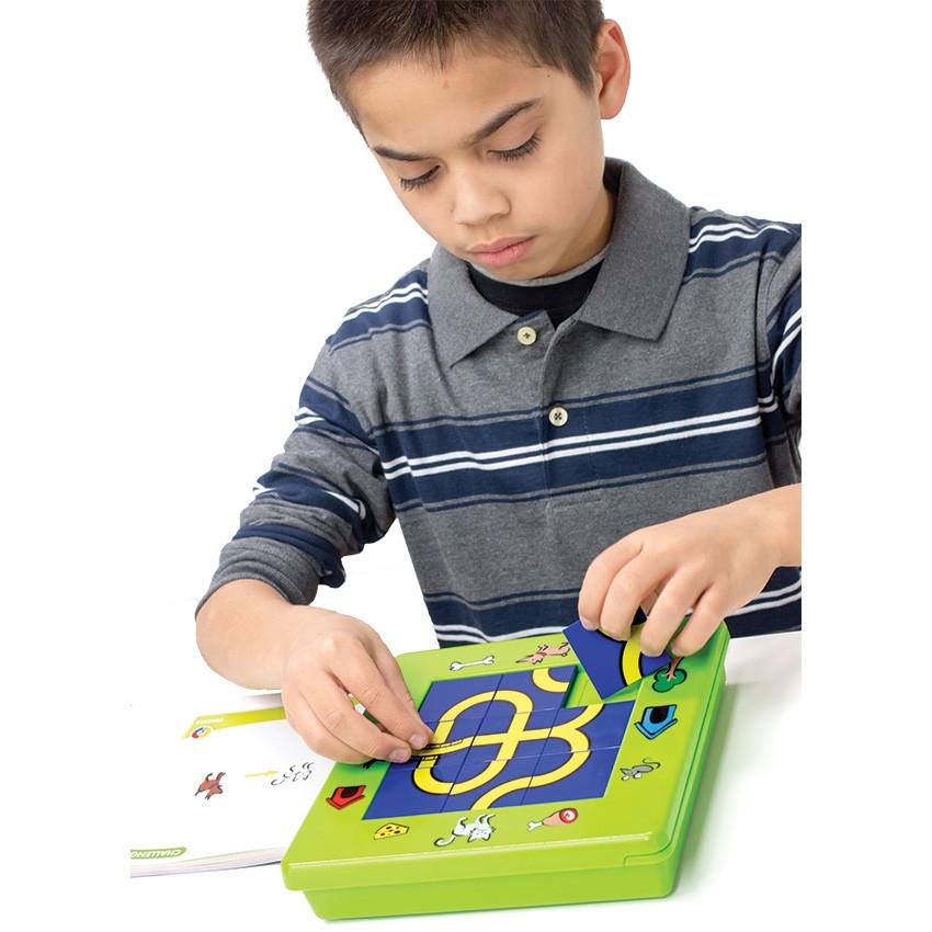 A young brunette boy playing with The Cat and Mouse game at a white table. The game board is bright green with illustrations around the board of pets, outside objects, pet snacks, and pet homes. There are blue and yellow square pathway pieces placed in the middle of the board, in a grid of 3 by 3. To the left is the instruction booklet, open to show a challenge. The boy is placing the last square piece into place on the board.