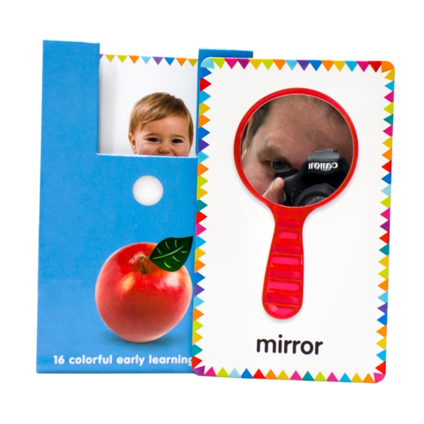 My First Touch and Feel Cards: First Words card pack open with a card propped up on the right of the pack. The packaging is blue with a red apple on the bottom. A card with a baby’s face can be seen inside the package. The card on the right shows a red hand mirror, with the reflection of the product photographer reflected in the mirrored surface. The background is white and there are colored triangle-shaped flags all around the border.