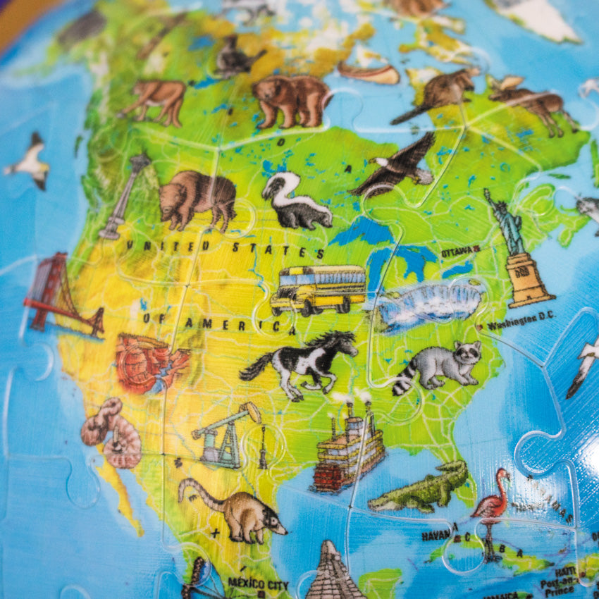 A close up of The Puzzleball Globe 180 piece set, put together. The 3 D globe portion in the image shows the U S A, with illustrations of animals, famous structures, and vehicles on top of the map.
