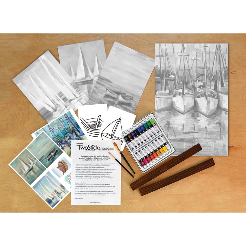 Two Stick Shadows Paint Kit, boats contents spread over a wood surface. 1 large canvas to the right shows 4 sailboats in a marina. Fanned out on the top-left are 3 medium canvases; 2 show sailboats, and 1 shows 2 row boats. Below, but on top, are 2 small canvases; 1 of a row boat, and 1 of a sailboat. Below, but on top, are 2 instruction sheets of the completed pieces and 1 page of text instructions. Over the top of instructions and large canvas are 2 paintbrushes, the 2 sticks frame, and set of 18 paints.