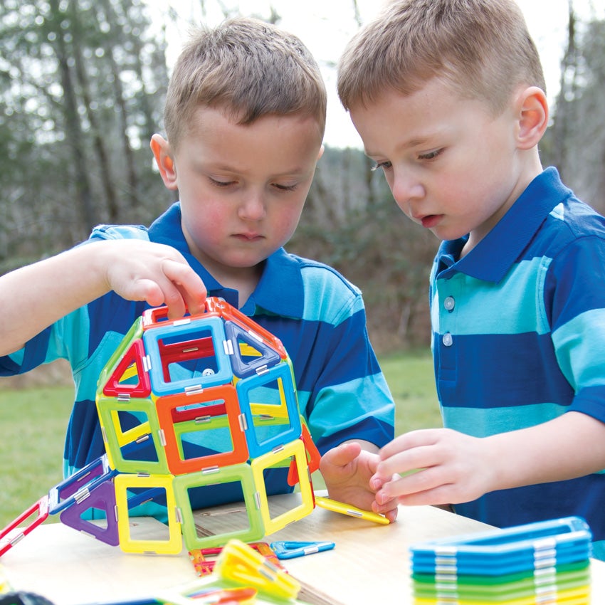 2 blonde boys in striped shirts are outside and are putting together a large shape, made up of the GeoSmart Educational Deluxe pieces on a picnic table. The pieces are geometrical shapes in squares and triangles and are a variety of different colors.  The object they are putting together is rounded and being held up by the boy on the left. The boy on the right is reaching out to grab a piece on the table. There are pieces in stacks and piles on the table.