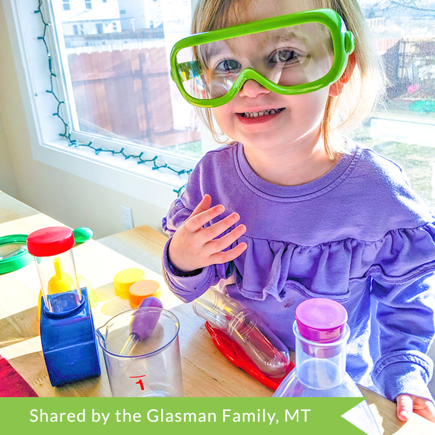 Customer photo of a blonde girl wearing safety goggles and a purple shirt. On the table in front of her are several items from the Primary Science Lab Kit. You can see a neighborhood in the windows behind her.