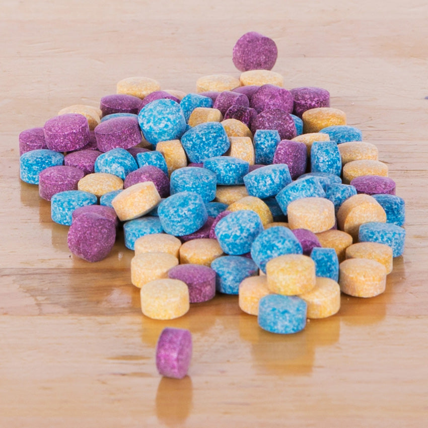 A close up shot of a pile of Color Fizzer tablets on top of a wood table. The colors of the tablets are purple, blue, and orange.