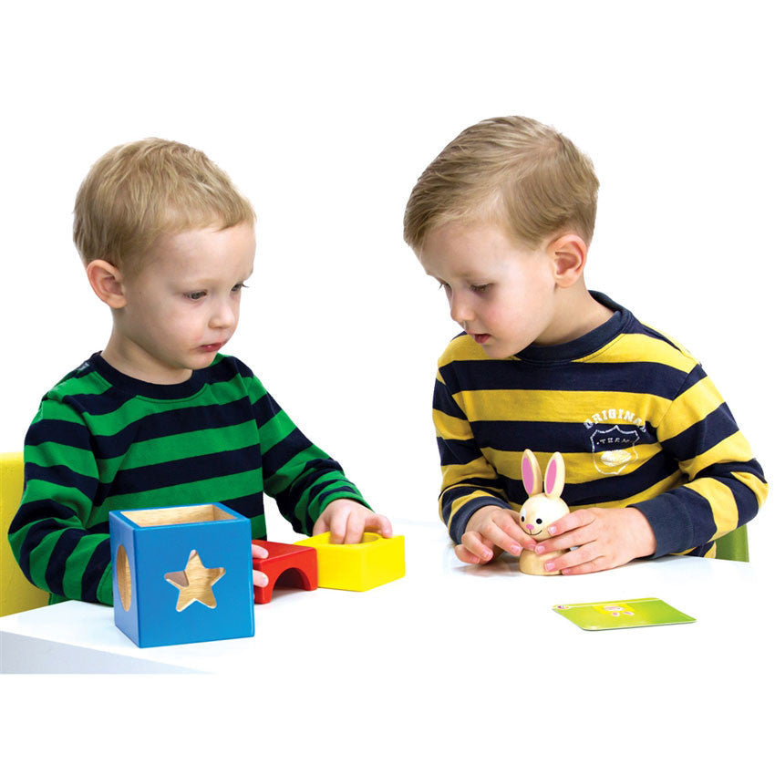 2 young blonde boys in striped shirts are playing with the Bunny Boo game. The boy on the left is placing the wood block pieces with his hand while looking at a challenge card, and the boy on the right is holding the bunny piece. The wood blocks are a hollow blue wood block that has shapes cut out on the sides, a yellow piece with a hollow circle through the middle, and a red piece that shows a half circle piece cut out of the middle.