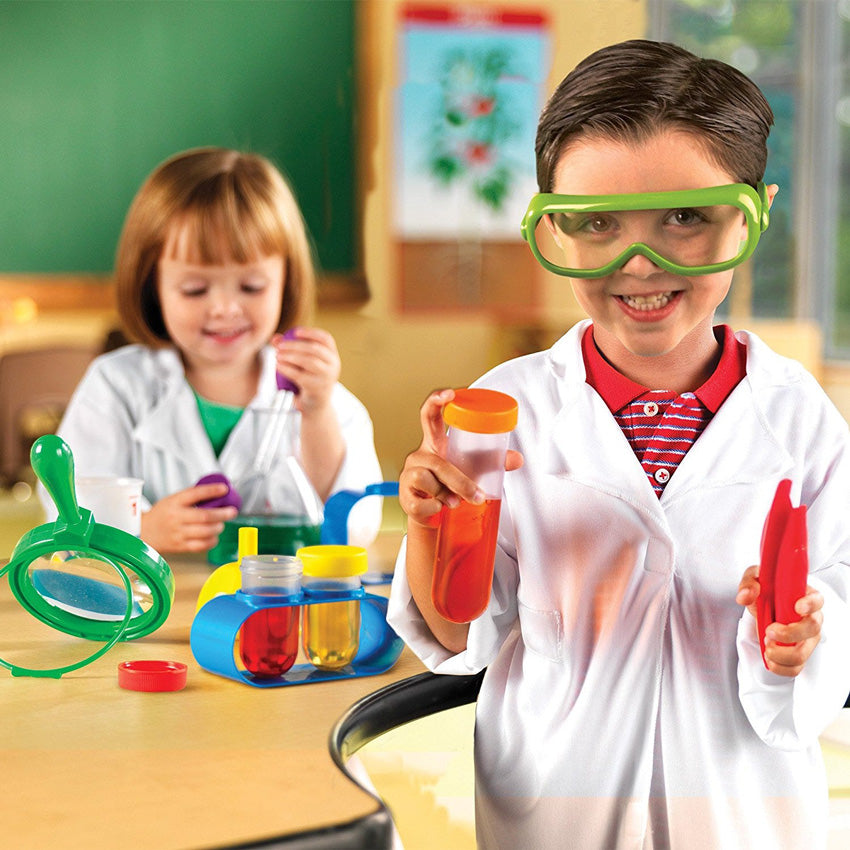 2 children using Primary Science Lab Set items. The boy in the front is holding a test tube with orange liquid and an orange lid in his right hand. He is holding red large tweezers in his right hand and green safety glasses with a white lab coat over a red striped shirt. Behind him is a table with science tools laid out with a girl playing with an eyedropper and green liquid. She is also wearing a lab coat over a green shirt.