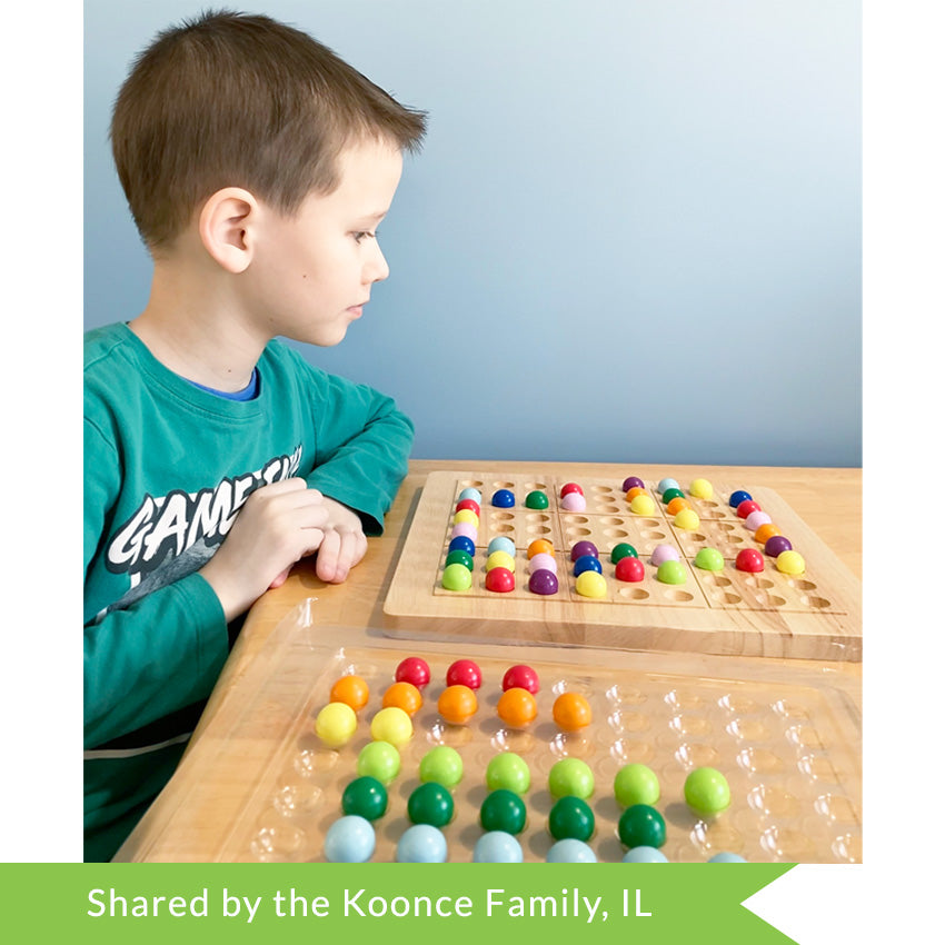 A customer photo of a brunette boy in the middle of playing ColorKu. He is looking down at the board in front of him on the table. The board in front of him has many pieces in place on top. On his right side is a clear tray for the ball pieces. The balls are in colored rows in the tray. The balls are red, orange, yellow, bright green, dark green, light blue, dark blue, light purple, and dark purple.