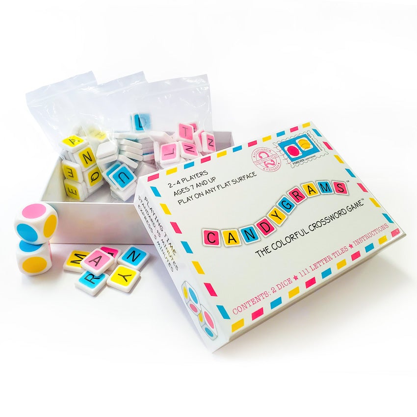 Candygrams game with the box open to show contents. The box lid, off to the right, is mainly white with the title in alternating colored tiles. Inside the box are 2 plastic bags filled with tiles; 1 bag with yellow tiles, 1 with blue tiles, and 1 with pink tiles. In front of the box bottom are 2 large dice with colored dots on each side, in pink, yellow, and blue. There is a small pile of colored tiles to the right of the dice.