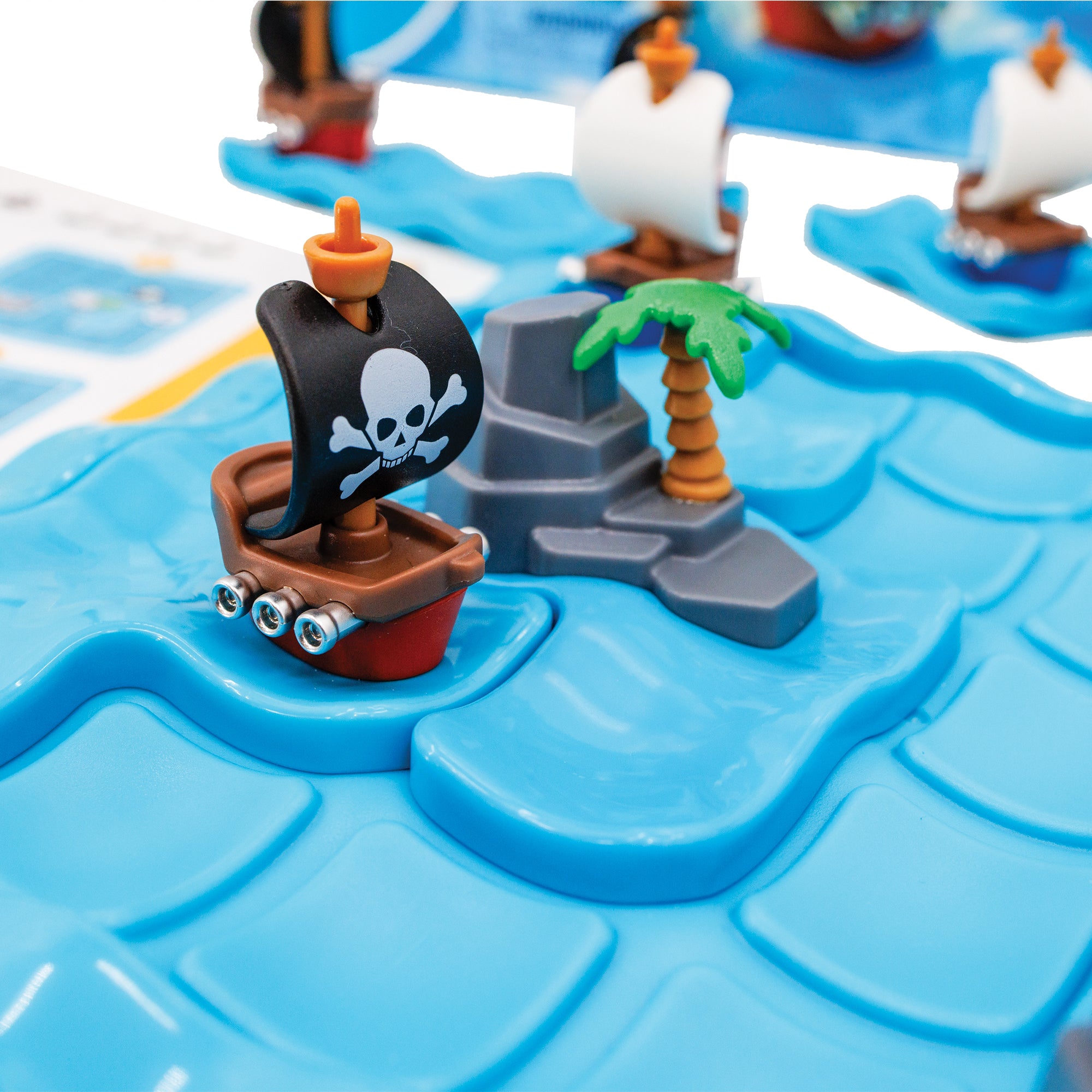 A close-up of the Pirates Crossfire game. The gameboard is blue and there are 2 pieces set in place on the board; one ship with a white skull on a black sail, and one rock piece with a palm tree. You can see pieces and the instruction book, out of focus, in the background.