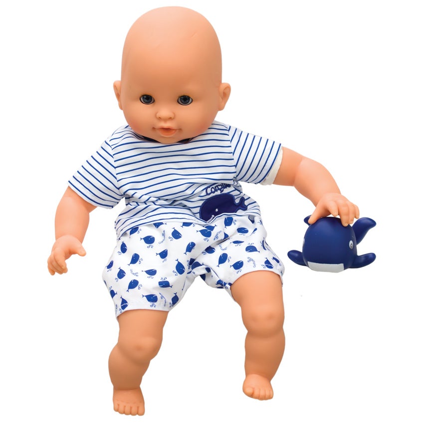 Corolle’s Bebe Marin sitting on a white background. The light skinned baby doll has blue blinkable eyes and is resting his hand on a rubber whale toy to his left. He is wearing shorts and a shirt. His shirt is white with blue stripes and a whale patch on his lower-left. His shorts are white with a blue whale pattern.