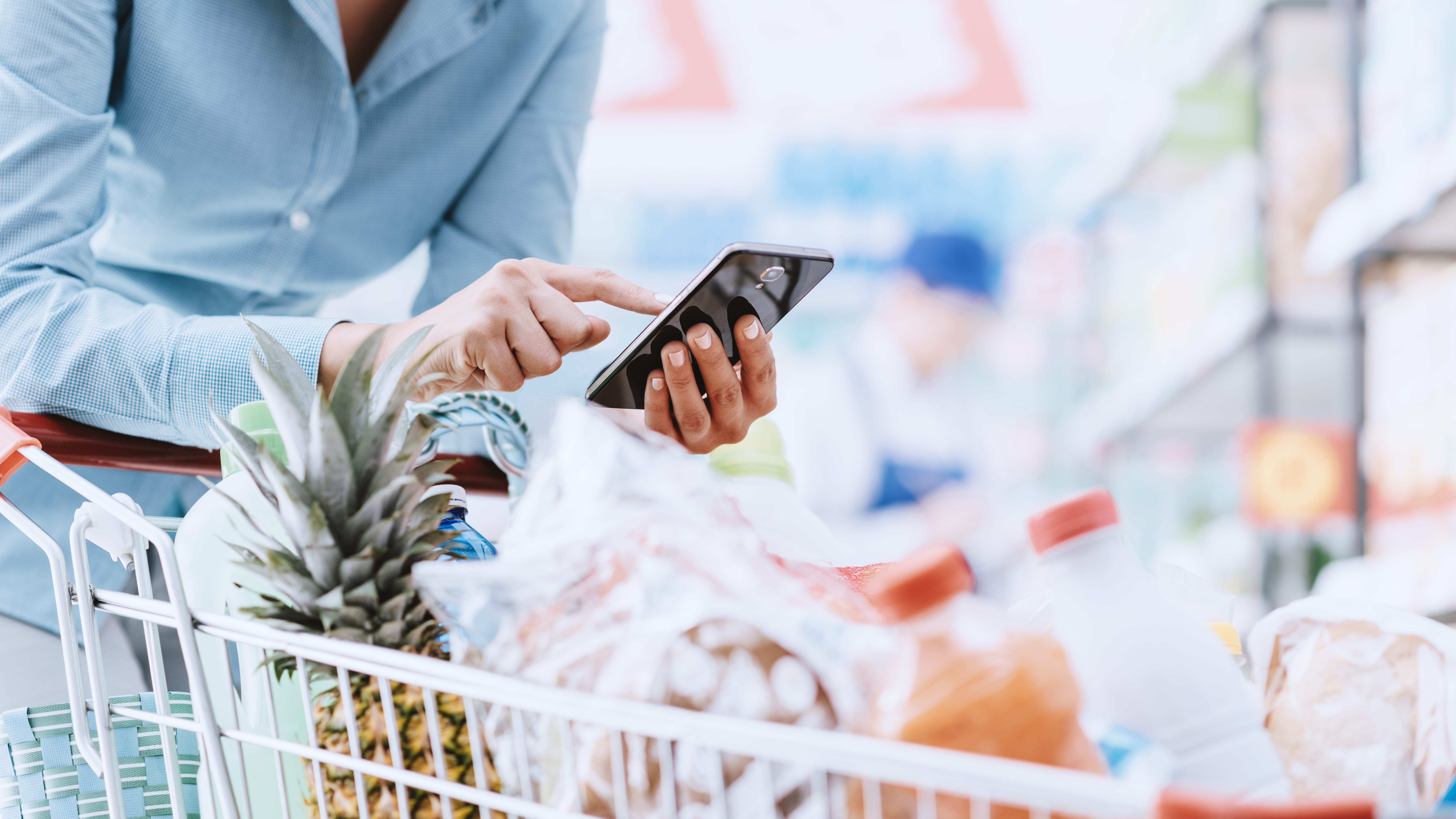 shopper in grocery store with a full cart and examining items on phone