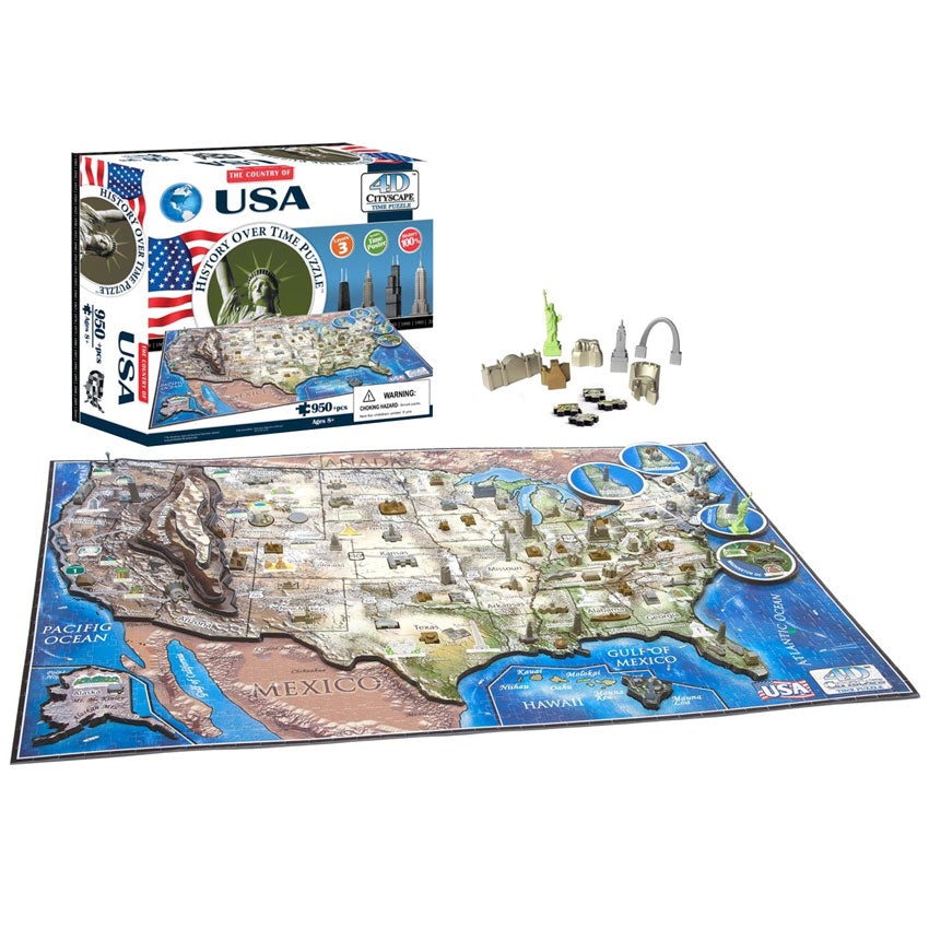 The U S A, History Over Time Puzzle box in front of a completed puzzle. The box is mainly white with a blue and white globe in the top-left corner. Across the middle is an American flag, The Statue of Liberty surrounded by a round border with the title inside, and a city scape. Below is the actual puzzle, put together, with several famous building pieces off to the side.